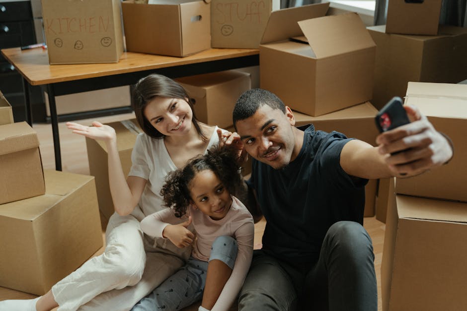 A woman with curly hair and light skin, dressed in beige trousers and a white shirt, is inside a modern, well-lit hallway of a house, carrying two large cardboard boxes, one placed on top of the other